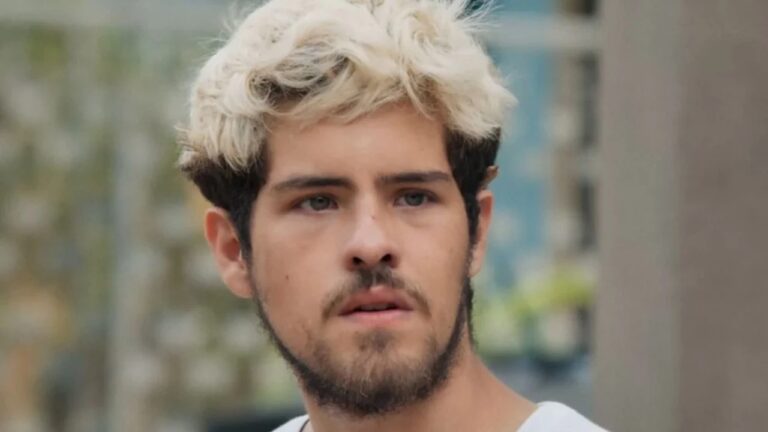 Homem com expressão séria e cabelos curtos descoloridos observa algo fora de cena, em um ambiente ao ar livre e urbano.