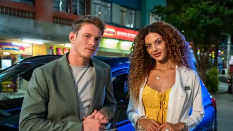 Homem e mulher posam lado a lado em frente a um carro estacionado à noite, com fachadas de comércios iluminadas ao fundo; ele usa blazer verde-claro e camiseta cinza, enquanto ela veste vestido amarelo estampado e jaleco branco, ambos olhando para a câmera.