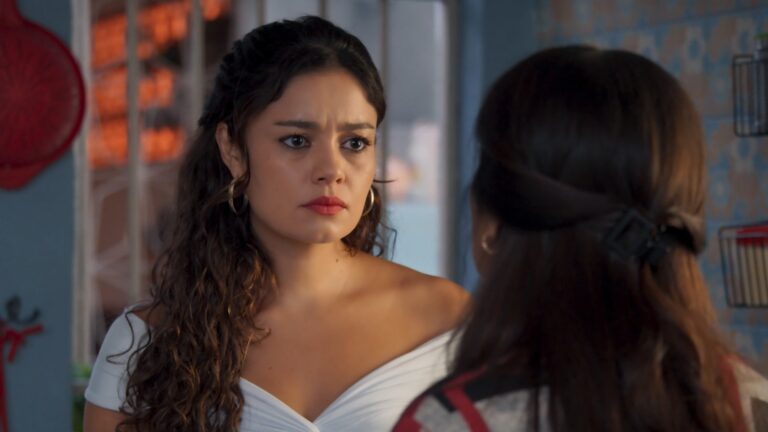 Cena de Três Graças mostra duas mulheres conversando em um ambiente doméstico. A mulher em primeiro plano, de blusa branca e expressão séria, olha atentamente para a outra, que aparece de costas. A iluminação natural e o cenário simples reforçam o tom dramático do momento