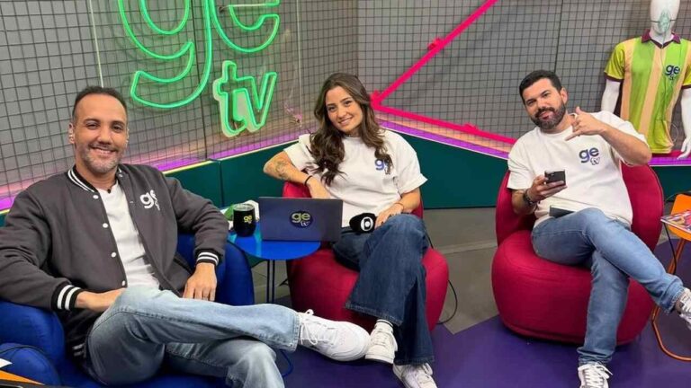 Três apresentadores sorriem sentados em poltronas coloridas no estúdio do programa “ge TV”, com o logotipo em neon ao fundo e clima descontraído.