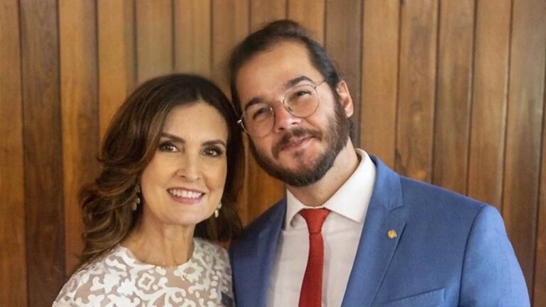 Casal posa sorridente diante de parede de madeira; a mulher usa vestido branco rendado e o homem veste terno azul com gravata vermelha.