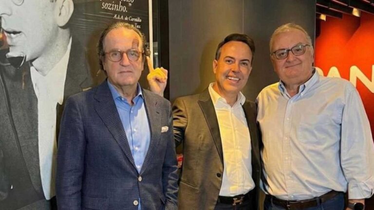 Três homens posam sorrindo em ambiente interno iluminado, dois deles usando paletó e o terceiro de camisa social azul clara, com fundo que exibe uma parede com fotografia em preto e branco e letras brancas parcialmente visíveis.