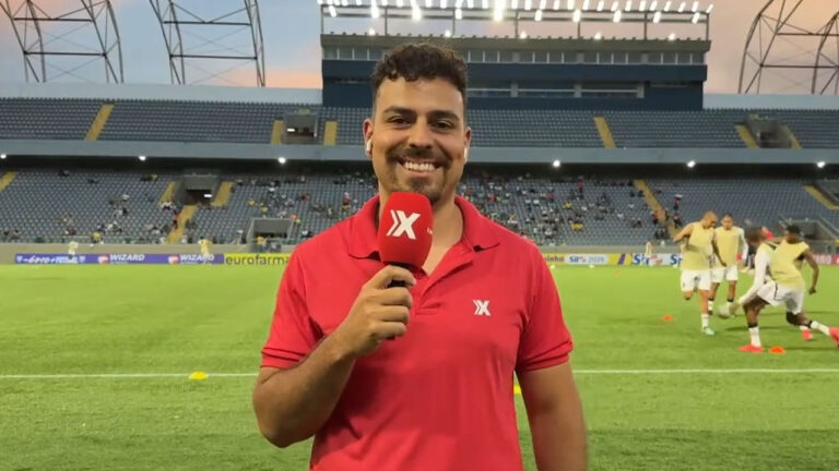 Repórter Tutti Batista sorridente segura microfone da Xsports com logotipo em destaque no gramado de estádio de futebol ao entardecer