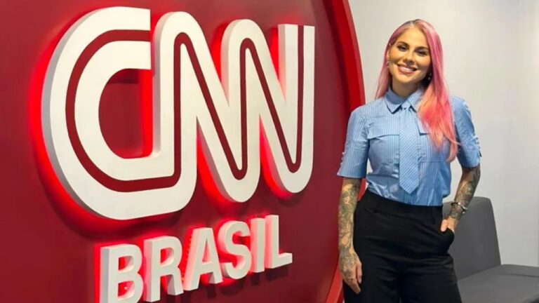 Mulher de cabelos longos e rosados sorri enquanto posa ao lado do logotipo da CNN Brasil em uma parede vermelha iluminada. Ela veste uma camisa azul clara com gravata curta combinando e calça preta, exibindo tatuagens nos braços. O ambiente é moderno e corporativo, com tons neutros e um sofá cinza ao fundo.