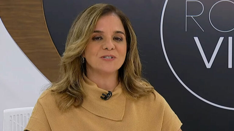 Vera Magalhães veste blusa mostarda de gola alta durante a apresentação do programa Roda Viva na TV Cultura com o logotipo da atração ao fundo