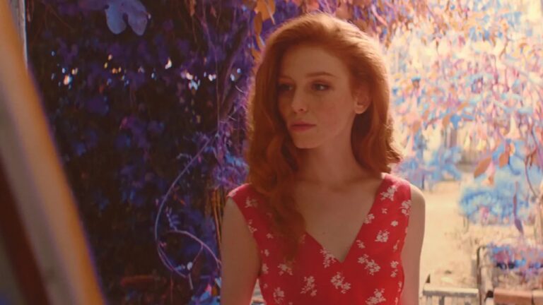 Mulher ruiva com vestido vermelho estampado observa alguém fora de cena em ambiente externo com folhagens coloridas ao fundo