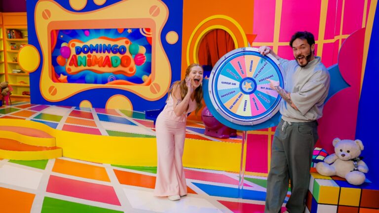 Dois apresentadores estão em um cenário colorido de programa infantil, com piso em blocos multicoloridos e fundo rosa, azul e amarelo. A mulher à esquerda veste macacão rosa claro e ri animada enquanto o homem à direita, de moletom cinza, gira uma roleta azul com prêmios escritos. Atrás deles há um grande letreiro colorido com o texto “Domingo Animado”.