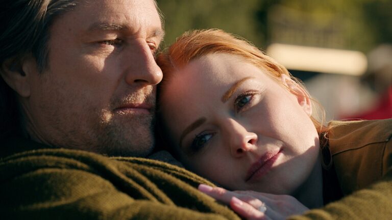 Homem e mulher estão deitados juntos em um momento íntimo e tranquilo ao ar livre. Ela apoia a cabeça no peito dele, com expressão serena e olhar distante, enquanto ele a observa de perto. A luz suave do sol ilumina o rosto dos dois, transmitindo um clima de carinho e contemplação.