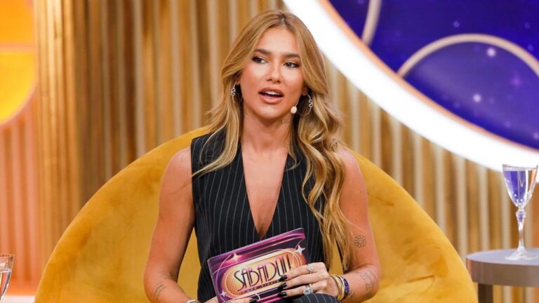 Mulher sentada em poltrona amarela apresenta o programa Sabadou com Virginia, conforme indica o cartão que segura. Ela usa colete preto com listras finas e microfone de lapela, enquanto fala em um estúdio iluminado com tons dourados e roxos ao fundo, criando um clima sofisticado e moderno