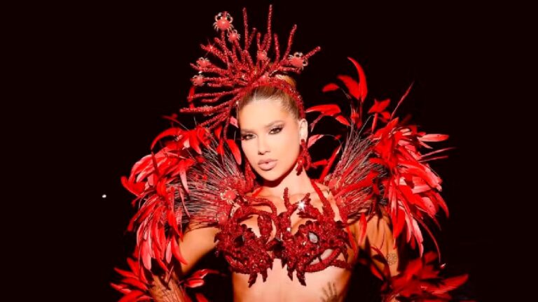 Mulher posa com expressão confiante usando uma fantasia exuberante em tons de vermelho, decorada com penas, pedras brilhantes e detalhes que lembram crustáceos. O adereço de cabeça é alto e ornamentado com estruturas semelhantes a corais, combinando com o corpo da fantasia. O fundo escuro destaca o brilho e a intensidade das cores do traje.