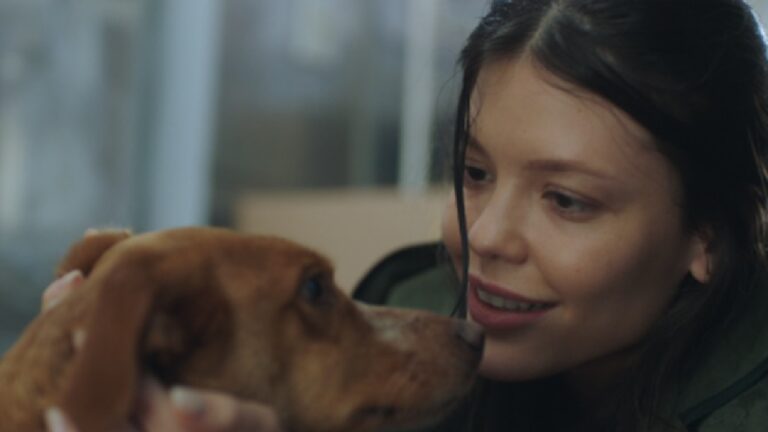 Mulher de cabelos escuros e expressão carinhosa segura o rosto de um cachorro marrom, olhando para ele de perto com um leve sorriso. O ambiente ao fundo é desfocado, sugerindo um local interno com luz suave.