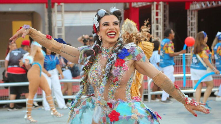 Vivian Cristinelle desfila sorrindo com fantasia colorida e tranças no cabelo durante apresentação de carnaval em avenida com público ao fundo