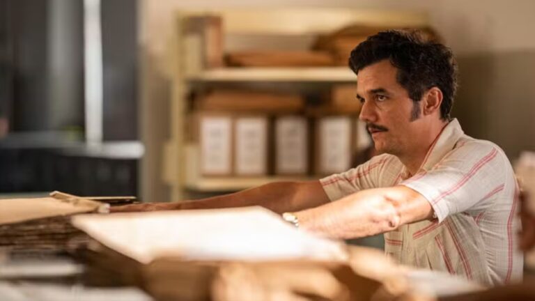 Homem com expressão séria está sentado em mesa coberta por pilhas de papéis, em ambiente de escritório iluminado; ele usa camisa clara com listras vermelhas e tem bigode.