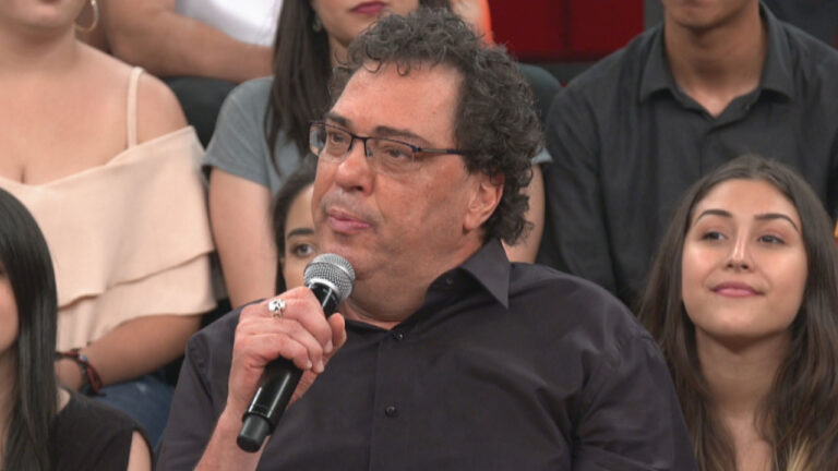 Walter Casagrande Júnior segura microfone e usa óculos e camisa preta durante participação em programa de TV com plateia ao fundo