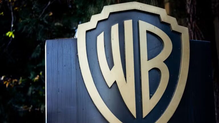 Placa metálica escura com o logotipo dourado da Warner Bros em destaque, formado por um escudo com as letras entrelaçadas “WB”. Ao fundo, há árvores e vegetação desfocadas, indicando que a placa está posicionada em uma área externa.