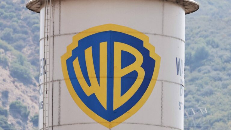 Torre de água branca exibe o logotipo azul e amarelo da Warner Bros em destaque no centro. À esquerda da estrutura há uma escada metálica vertical, e ao fundo aparece uma paisagem de colinas cobertas por vegetação.