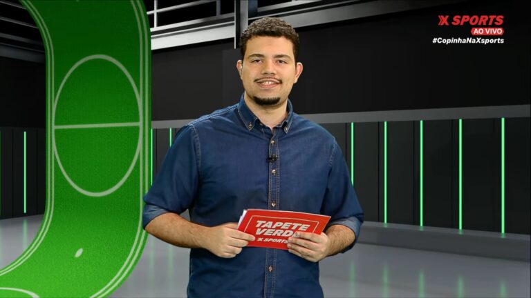 Zé Neto, apresentador do canal XSports, aparece em estúdio moderno com fundo escuro e detalhes em verde, segurando um cartão vermelho com o título Tapete Verde. Ele veste camisa jeans e sorri para a câmera durante transmissão ao vivo do programa esportivo
