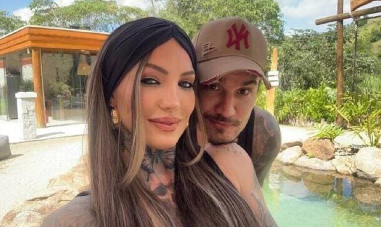 Mulher e homem posam juntos sorrindo levemente em frente a uma piscina ao ar livre, cercada por plantas e pedras. A mulher, com cabelos longos e lisos, usa uma faixa preta na cabeça e brincos dourados, enquanto o homem, de boné bege com logotipo vermelho, está atrás dela, com o braço apoiado em seu ombro. Ao fundo há uma construção moderna de madeira e vidro em meio a área arborizada.