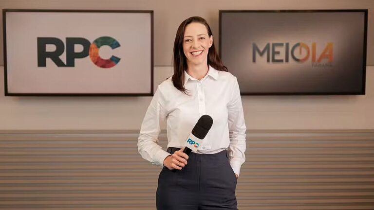 Repórter sorri para a câmera segurando um microfone com a logomarca “RPC”, em ambiente interno corporativo. Ao fundo, dois monitores exibem os logos “RPC” e “Meio Dia Paraná”, indicando gravação ou apresentação de telejornal.