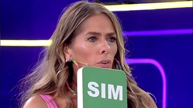 Mulher loira aparece em um estúdio de TV com iluminação em tons de roxo e azul ao fundo. Ela usa um vestido rosa e brincos coloridos, além de um microfone preso ao rosto. Na mão, segura uma placa verde com a palavra “SIM” escrita em branco. Sua expressão é séria e concentrada, como se estivesse reagindo a algo durante o programa.