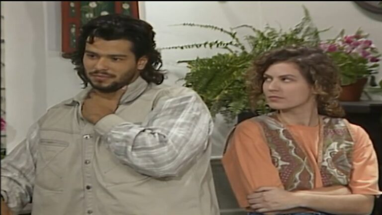 Um homem e uma mulher estão lado a lado em um ambiente interno, com plantas e quadros ao fundo. O homem, de cabelos médios e barba, veste camisa clara com colete e segura a gola próxima ao pescoço, olhando à frente com expressão séria. A mulher, de cabelos curtos e cacheados, usa blusa alaranjada com colete estampado, mantém os braços cruzados e olha para ele com semblante desconfiado ou irritado. A cena sugere um momento de tensão ou discussão.