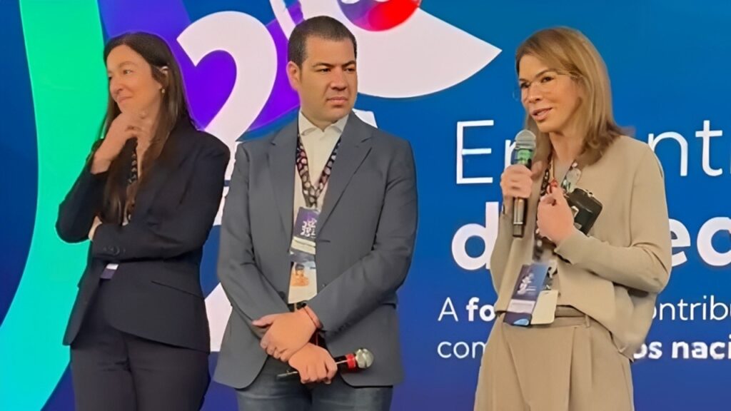 Alessandra Ribeiro, Daniel Abravanel e Daniela Beyruti estão em um palco durante um evento corporativo do SBT. As mulheres e o homem usam crachás e roupas formais; Daniela segura um microfone e parece estar falando ao público, enquanto os outros dois observam