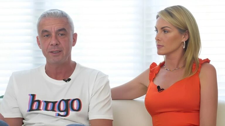 Homem de cabelos grisalhos, usando camiseta branca, fala durante entrevista enquanto mulher de vestido laranja o observa; ambos estão sentados em ambiente interno iluminado, com microfones de lapela visíveis.