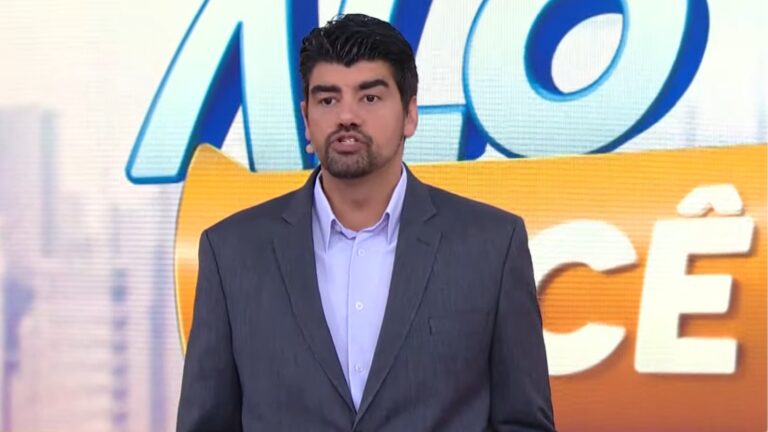 André Azeredo, de terno cinza e camisa clara aparece em estúdio de TV, falando diante de um fundo colorido com o logotipo do programa Alô Você. Ele usa microfone de lapela e mantém expressão séria enquanto apresenta