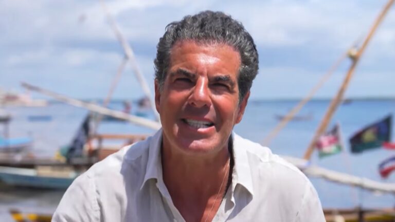 Alvaro Garnero, usando uma camisa clara, aparece ao ar livre em um cenário litorâneo ensolarado no Quênia, com barcos e bandeiras coloridas ao fundo. Ele está apresentando o 50 por 1, programa de viagens exibido dentro do Domingo Espetacular, da Record