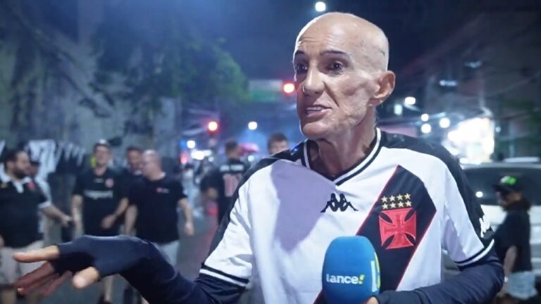 Homem careca veste camisa do Vasco da Gama e fala com repórter durante entrevista em rua à noite, com torcedores e viaturas ao fundo sob iluminação urbana.
