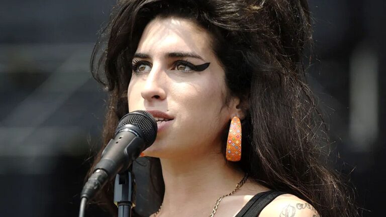 Cantora de cabelos longos e escuros se apresenta ao ar livre, cantando ao microfone. Ela usa delineado preto marcante estilo retrô, brincos grandes alaranjados e colar fino. A imagem é um close no rosto da artista, que tem expressão intensa e concentrada durante a performance.