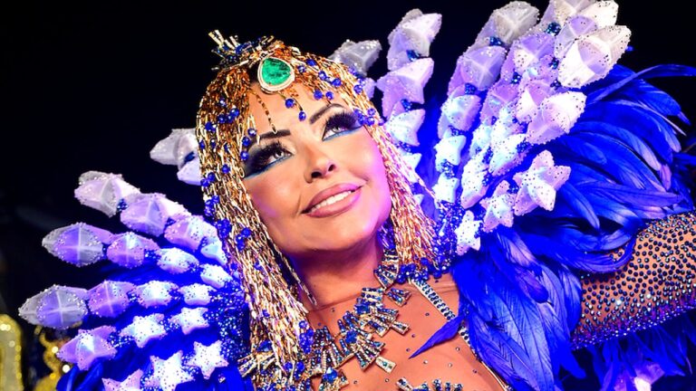 Ana Beatriz Godoi sorri usando fantasia de Carnaval luxuosa com adereço de cabeça dourado, pedrarias azuis, colar ornamental e grandes asas de penas