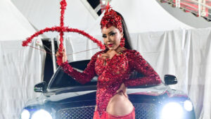 Ana Beatriz Godoi posa com fantasia vermelha de brilhos e pedrarias segurando um arco decorativo em frente a um carro de luxo com faróis acesos à noite