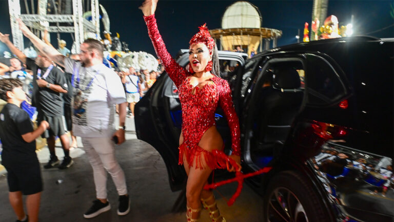 Ana Beatriz Godoi de fantasia vermelha brilhante com franjas e adereço de cabeça desce de carro preto e acena no sambódromo durante o Carnaval à noite