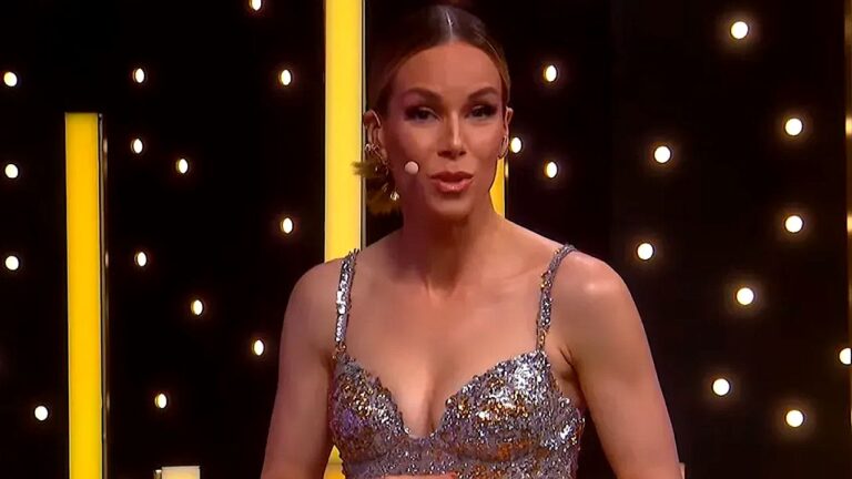 Apresentadora aparece em estúdio iluminado, usando vestido prateado com brilho e alças finas, além de microfone preso ao rosto. Ao fundo, há painéis verticais amarelos e luzes circulares que remetem a um palco de programa de entretenimento. Ela olha para a câmera com expressão confiante, como se estivesse conduzindo a atração ao vivo.