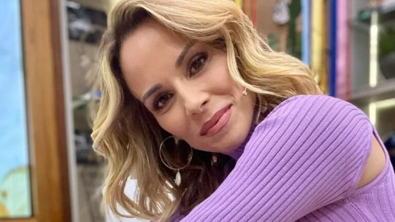 Apresentadora posa sorrindo com blusa roxa e cabelo loiro ondulado em estúdio de programa de TV durante gravação