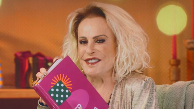 Ana Maria Braga sorri segurando livro rosa com logotipo colorido, usa blusa metalizada dourada, possui cabelos loiros curtos e fundo com luzes desfocadas