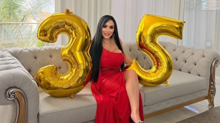 Ana Paula Oliveira de cabelos longos e pretos usa vestido vermelho longo e posa sentada em sofá cinza entre balões dourados que formam o número 35