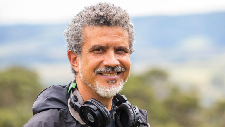 Diretor André Câmara sorri de barba grisalha e fones de ouvido no pescoço em plano médio com fundo de paisagem natural desfocada durante o dia
