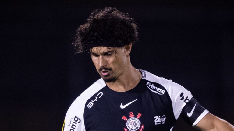 André Ramalho olha para baixo com expressão séria, veste camisa preta e branca do Corinthians com diversos patrocinadores, usa faixa preta na testa, tem cabelos cacheados e fundo escuro