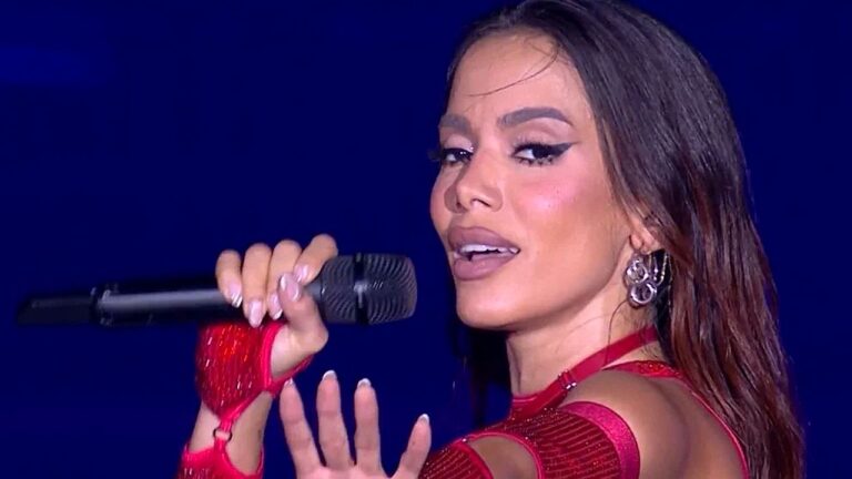 Cantora se apresenta no palco segurando um microfone, usando figurino vermelho brilhante com detalhes vazados e luvas combinando. Com maquiagem marcante e delineado destacado, ela olha para o público enquanto canta, diante de fundo iluminado em azul, sugerindo show ou apresentação televisiva.
