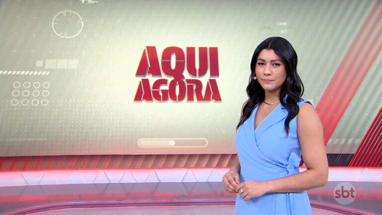 Daniele Brandi, mulher de cabelos longos e escuros, apresenta telejornal em estúdio, vestindo vestido azul-claro sem mangas; ao fundo, telão com a inscrição Aqui Agora em letras vermelhas e logotipo do SBT no canto da tela.