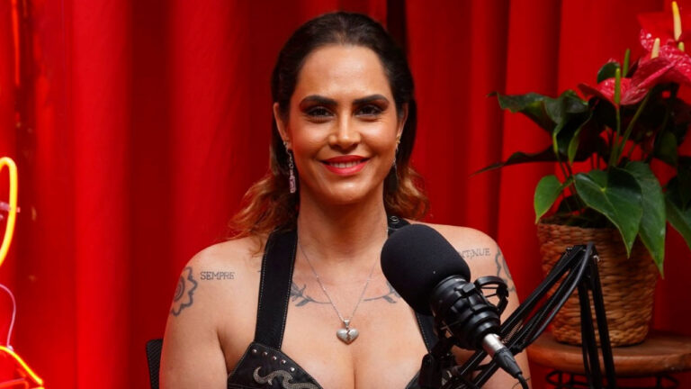 Aritana Maroni sorridente de blusa preta com tachas, colar de coração e tatuagens nos ombros, em frente a um microfone de podcast com fundo vermelho