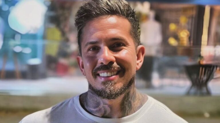 Arthur O Urso sorri de frente vestindo camiseta branca, exibe barba aparada e tatuagens que cobrem o pescoço em cenário urbano com luzes desfocadas ao fundo