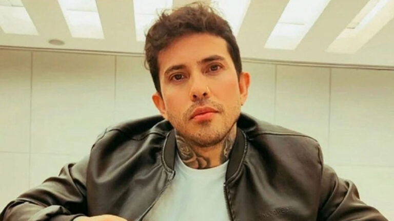 Influenciador Arthur O Urso de jaqueta de couro preta e camiseta branca exibe tatuagens no pescoço enquanto posa em ambiente com teto iluminado ao fundo