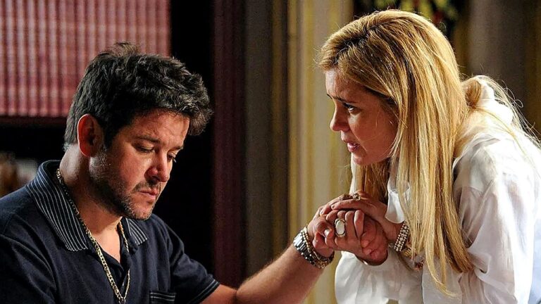 Um homem e uma mulher estão próximos, em um ambiente interno com estantes ao fundo. O homem, com cabelo escuro curto e barba por fazer, veste uma camisa escura e acessórios como corrente e pulseiras. Ele está sentado, olhando para baixo com expressão abatida ou pensativa. A mulher, loira e usando uma blusa branca, inclina-se em direção a ele e segura sua mão com as duas mãos, demonstrando apoio e preocupação. A cena transmite um momento íntimo e emocional, sugerindo consolo ou uma conversa séria entre os dois.