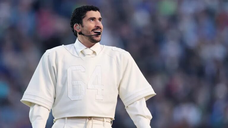 Cantor Bad Bunny usa figurino branco com número 64 e microfone durante apresentação em grande estádio com público ao fundo