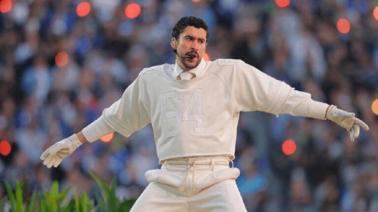 Cantor se apresenta em um grande estádio lotado, usando roupa branca com o número “64” estampado no peito. Ele abre os braços durante a performance e canta com microfone preso ao rosto, enquanto a plateia aparece desfocada ao fundo, com luzes e celulares iluminando o ambiente.