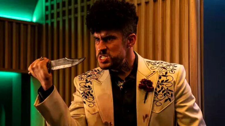 Homem segura uma faca com expressão de raiva em ambiente interno iluminado por luzes verdes e amarelas. Ele veste terno claro com bordados escuros e flor na lapela, além de camisa preta, e parece estar em momento de confronto. O cenário moderno, com painéis de madeira ao fundo, sugere cena tensa de filme ou série dramática.