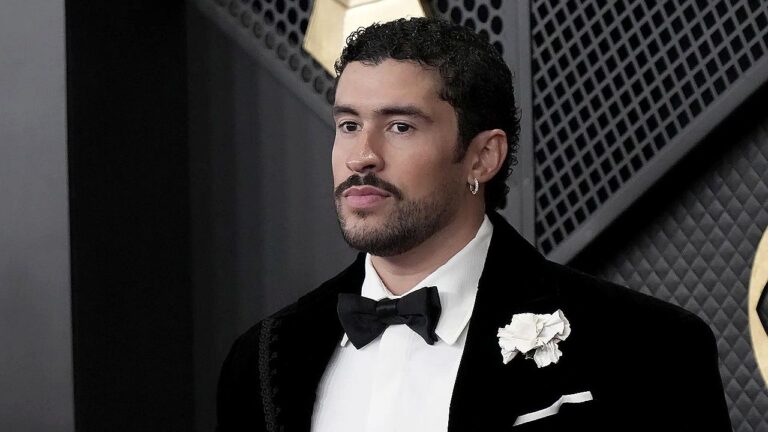 Homem veste smoking preto com gravata borboleta e flor branca na lapela, em evento formal. Ele tem barba aparada e brinco discreto, posando diante de fundo elegante com painéis escuros e iluminação suave, sugerindo cerimônia ou premiação.